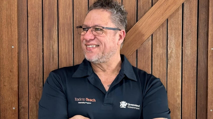 A person with glasses and short hair smiles while crossing their arms, wearing a "Trade to Teach" shirt with a Queensland Government logo, standing in front of a wooden backdrop.
