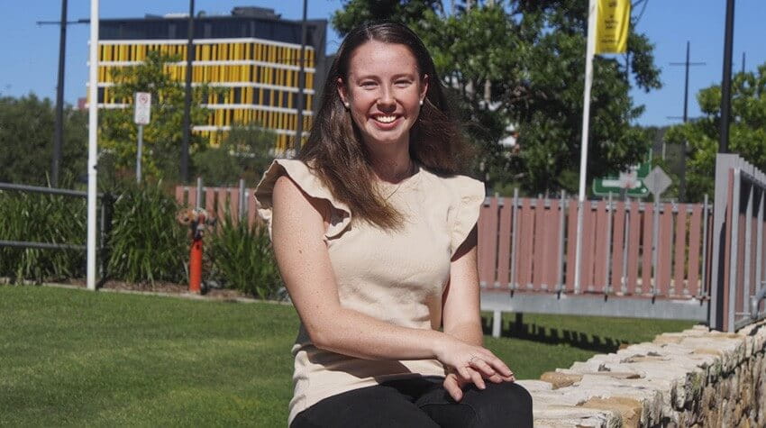 Emma is sitting on outside on a sandstone bench, smiling at the camera.