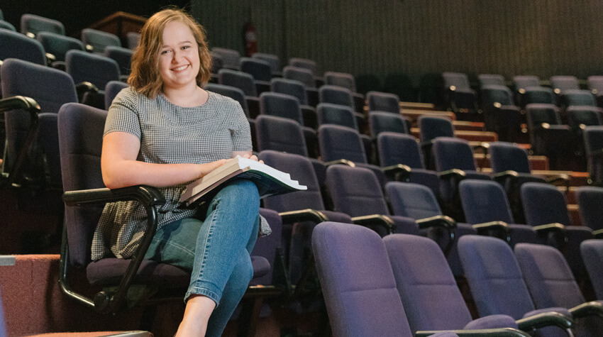 Emma is sitting in a lecture lecture theatre with her text books, smiling at the camera.