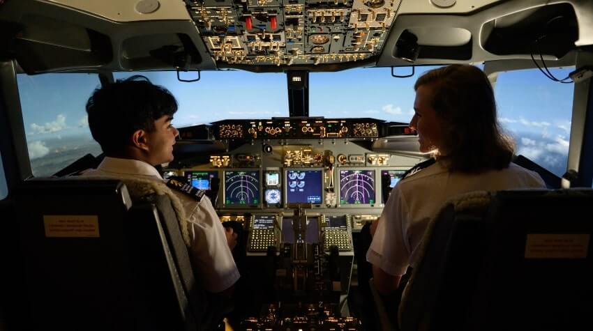 Two pilots sit in the cockpit of an aircraft, facing the control panels and looking at each other while flying above the clouds.