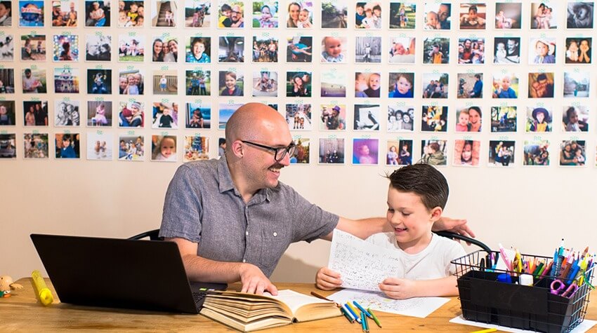 Father sits with son at table with laptop, book, pens and paper.