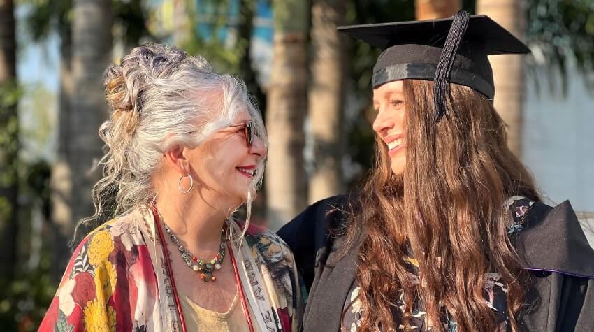 An older woman and a young woman in a graduation cap smile at each other outdoors.