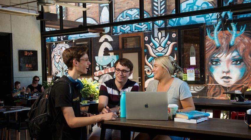 Three individuals engaging in a discussion at a café with laptops and books on the table, and a colorful mural in the background.
