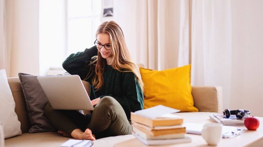 Woman sitting comfortably on a couch using a laptop, with books and a camera beside her.