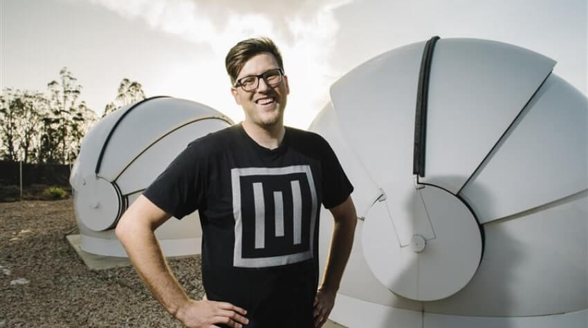 A person wearing glasses and a T-shirt with a geometric design stands smiling in front of two large, dome-shaped structures outdoors.