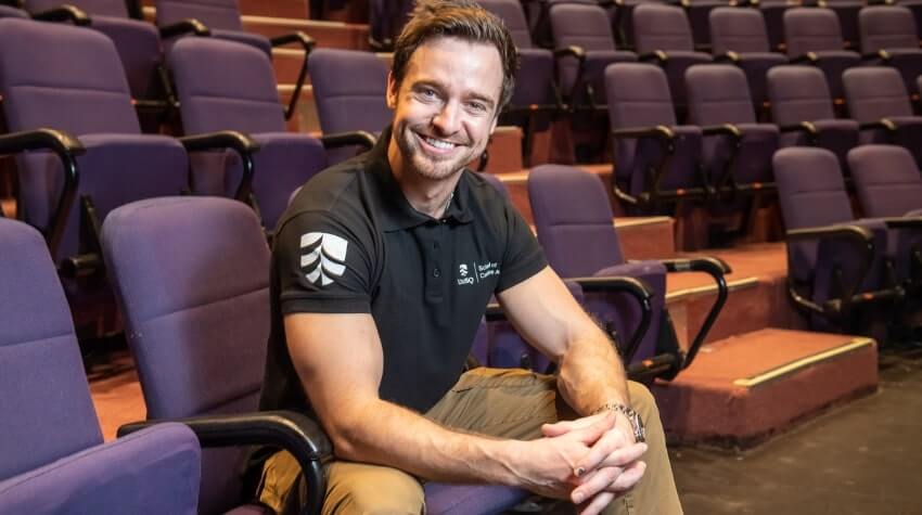A person smiling and seated in an empty theater with purple seats, wearing a black polo shirt and khaki pants.
