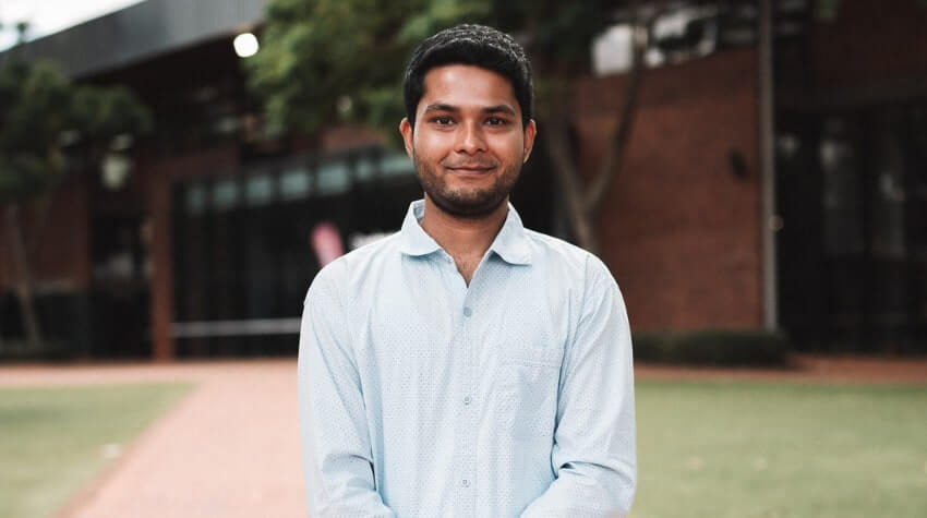 A man in a light blue shirt stands outside on a path, with grass and a brick building in the background.