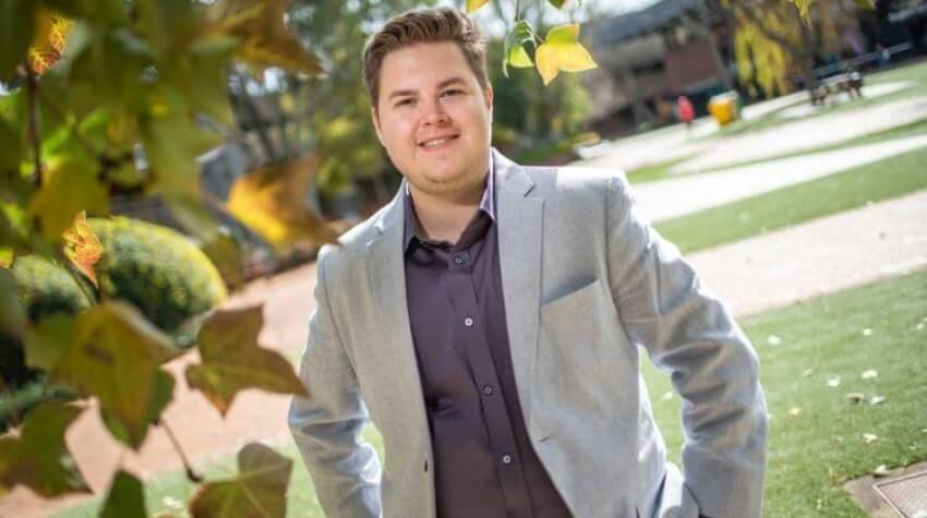 Person in a light gray blazer stands outside in a park-like area with green grass and trees.