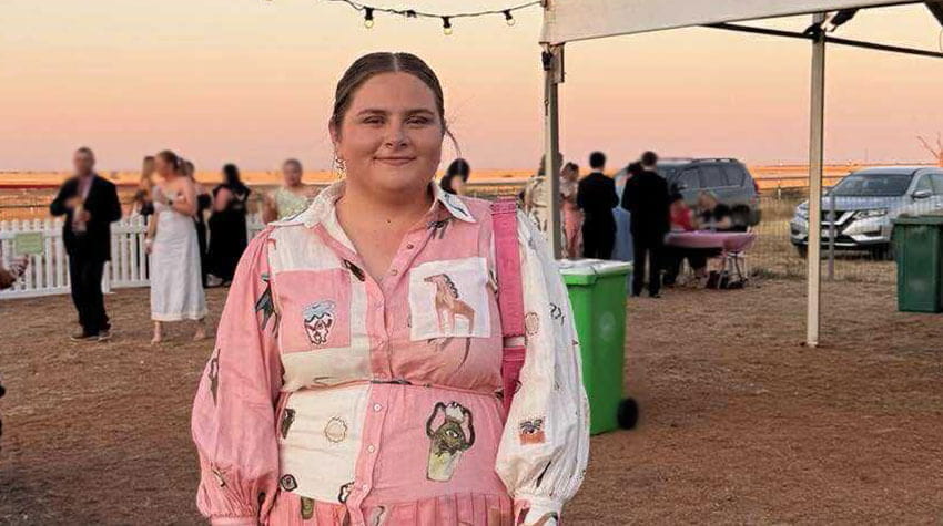 A woman in a pink and white patterned dress stands outdoors at an event, with people and cars in the background at sunset.