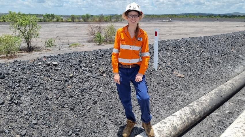 Person in orange safety gear and helmet stands on gravel near large pipes in an open area. Trees and clouds are visible in the background.