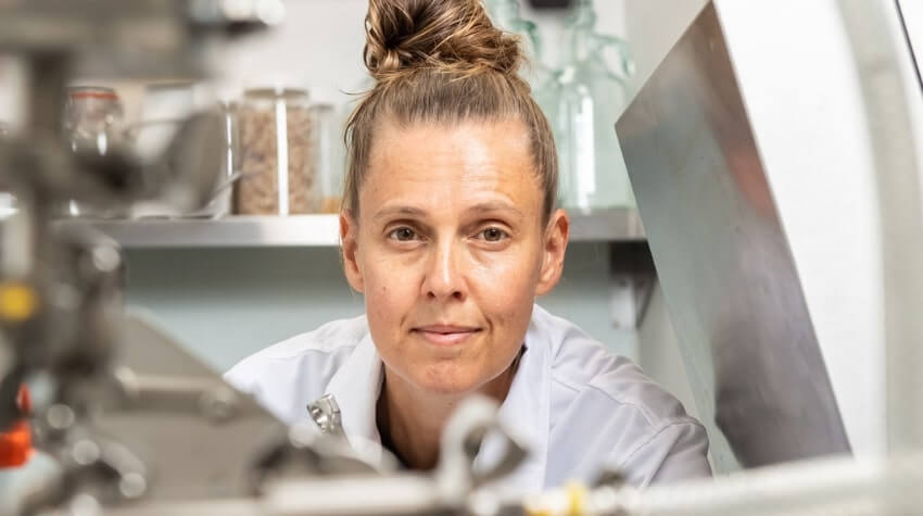 A woman in a lab coat, with hair in a bun, looks at the camera while standing in a laboratory setting.