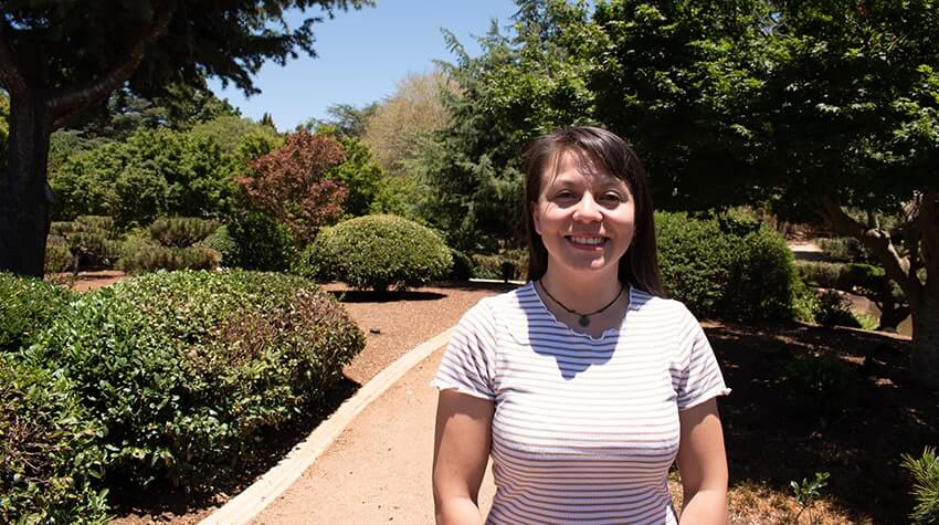 Maria is standing in the Japanese Garden, UniSQ Toowoomba, smiling at the camera.