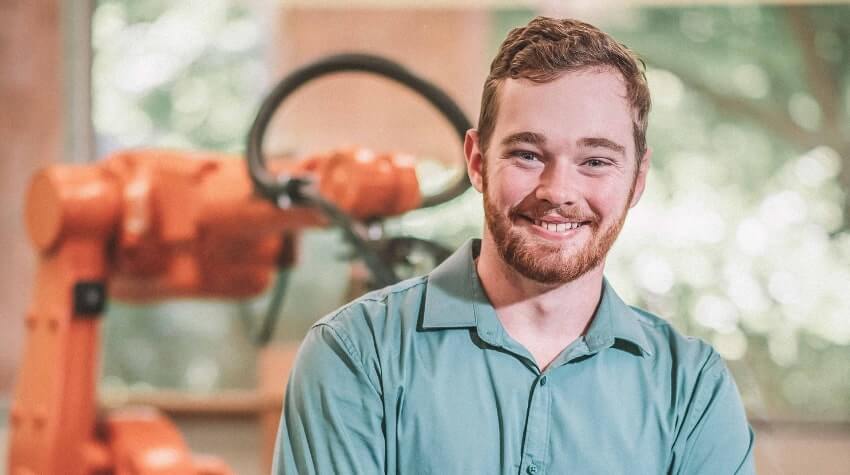 A smiling person in a green shirt with a robotic arm in the background.