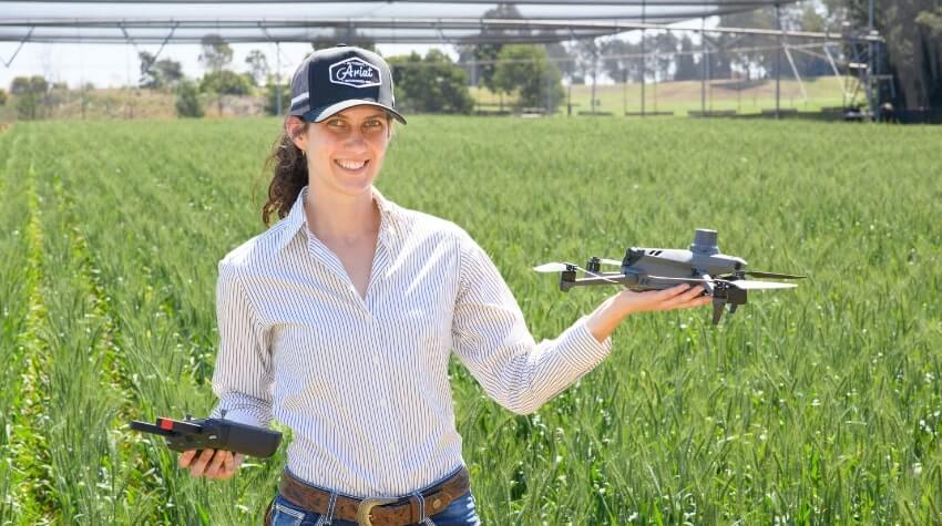 Person in a striped shirt and cap holds a drone and remote control in a field, with agricultural equipment in the background.