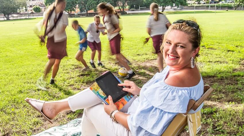 Woman relaxing with tablet in the foreground with blurred children playing in the background at a park.
