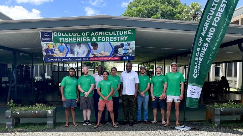 A group of people in green shirts stand with another individual in front of a building with a "College of Agriculture, Fisheries & Forestry" banner and flag.