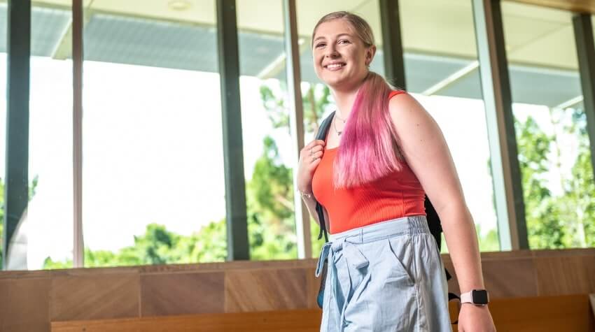 A smiling young woman with a backpack standing indoors by a window.