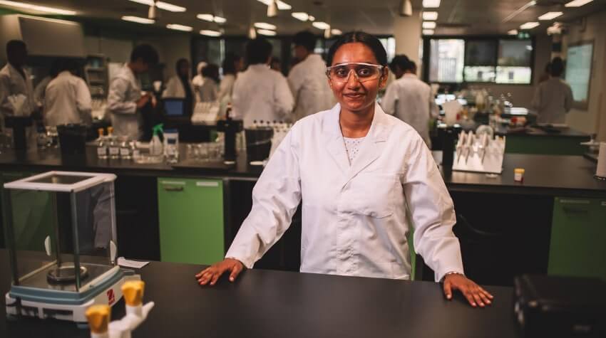 A scientist in a lab coat and safety glasses stands at a lab bench. Other people in lab coats work in the background.
