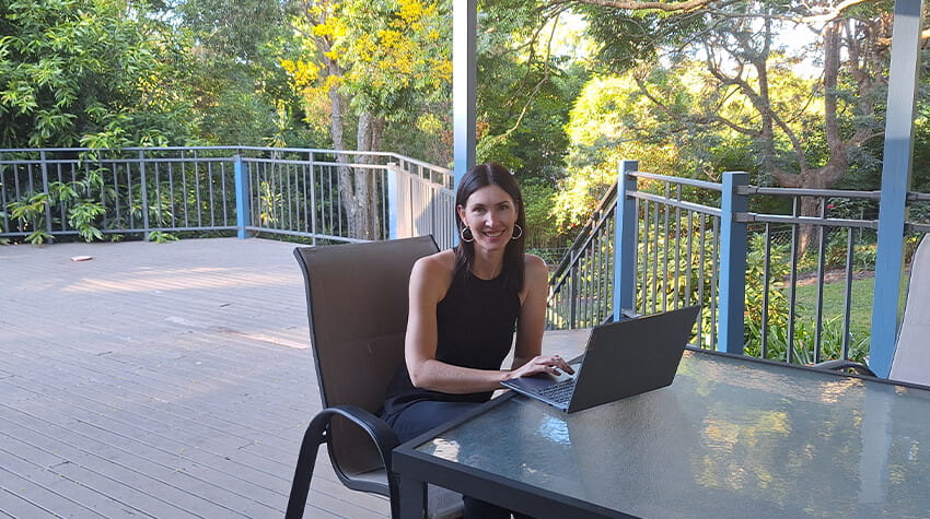 A woman working on a laptop at an outdoor table surrounded by greenery.