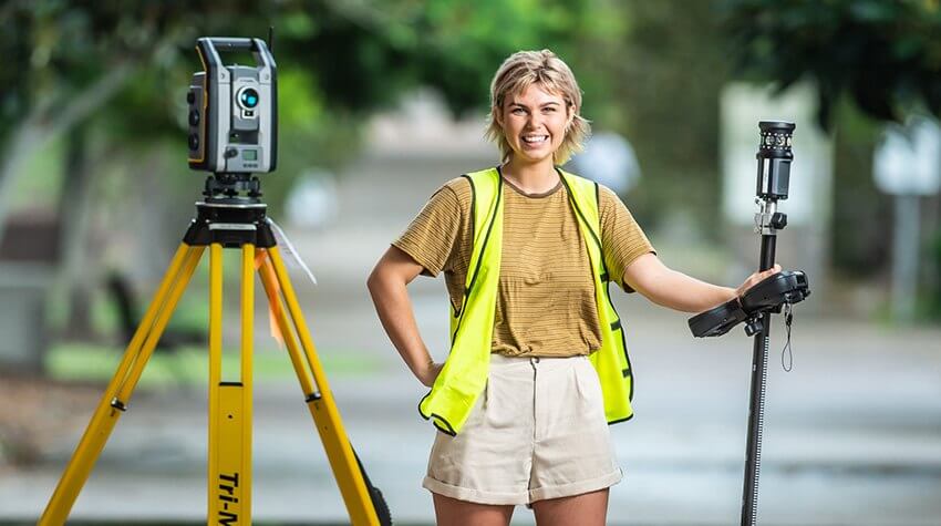 Rebecca holding and standing next to surveying equipment, smiling into the camera.