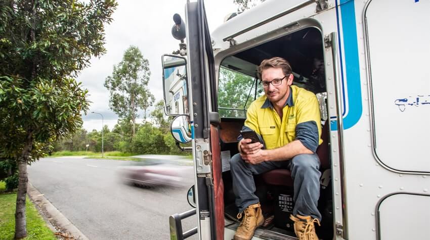 A male truck driver in high-visibility clothing taking a break and using his smartphone by the open door of his semi-truck.