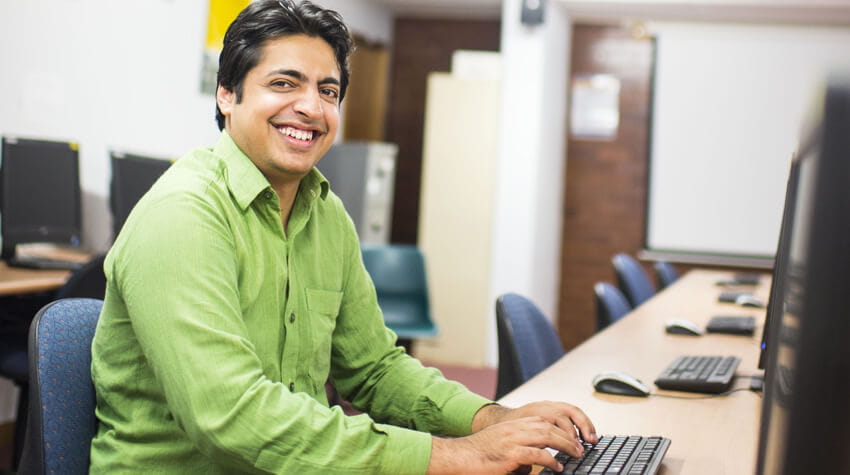 Man smiling at the camera while working on a computer in an office environment.