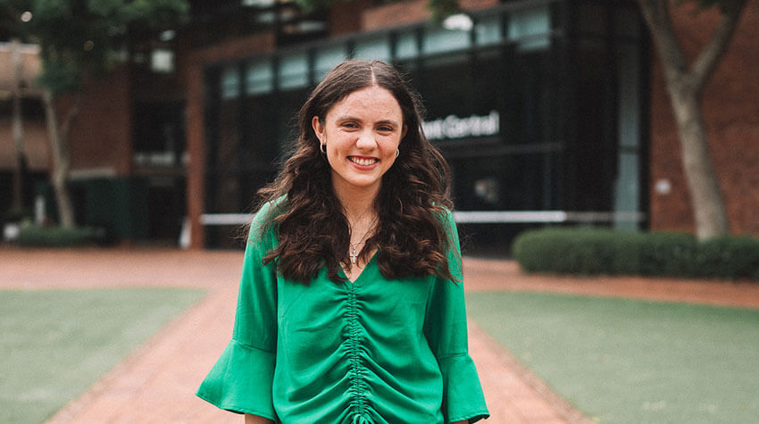 A woman with long brown hair, wearing a green blouse, stands smiling outdoors on a brick walkway with trees and a building in the background.