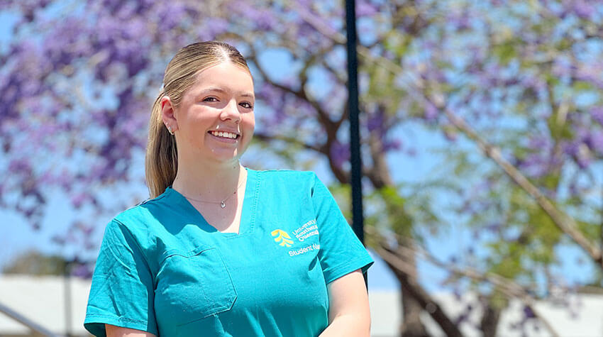 A woman in teal medical scrubs stands outdoors, smiling, with trees and purple flowers in the background.