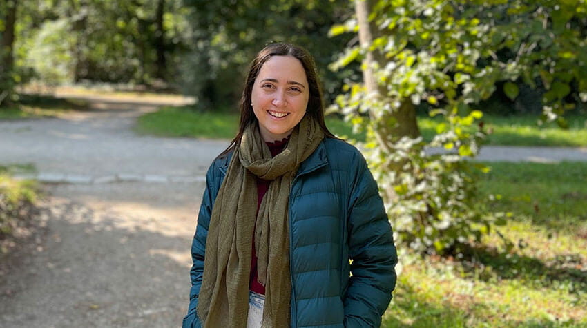 A woman wearing a teal jacket and olive scarf stands on a sunlit path in a wooded park, smiling at the camera.