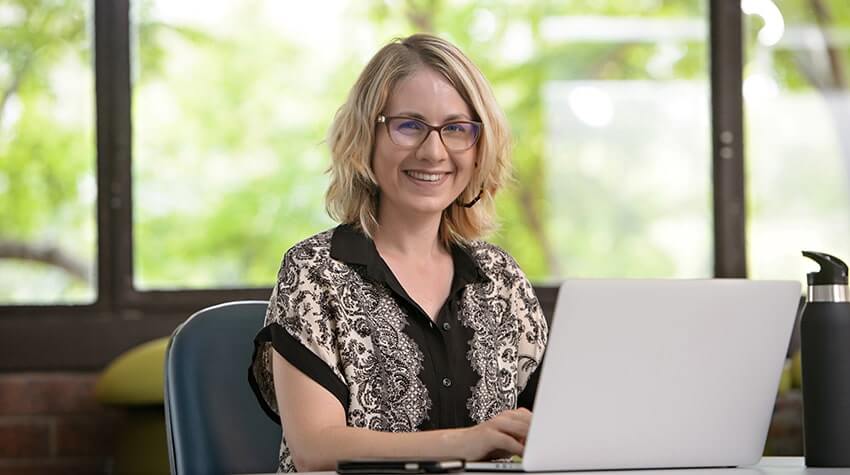 Woman with glasses smiling at the camera while working on a laptop in an office setting.