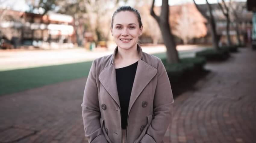A person stands outdoors on a brick path, wearing a coat, with trees and buildings in the background.