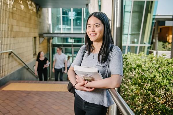 Student waiting outside university campus building holding book.